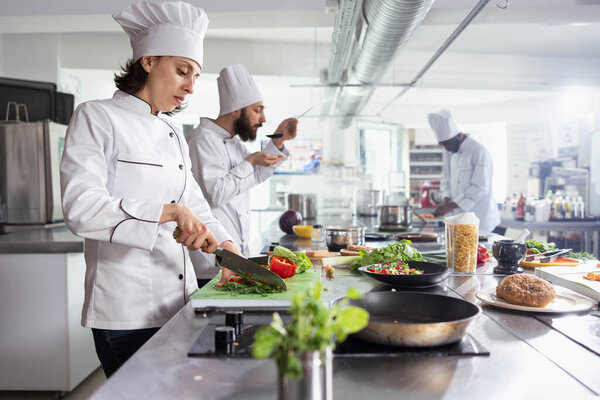 Focused female chef cutting small pieces of flavory herbs for a dish on the stove, mincing rosemary or basil on the cutting board. Woman cook using greenery and condiments for tasty meal.
