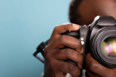 Stüdyoda siyah adamın DSLR kamerasını mavi arka planda tuttuğu aşırı yakın plan. Erkek fotoğrafçı, kamera kullanım tekniklerini objektife özel olarak odaklıyor, karmaşık detayları vurguluyor.