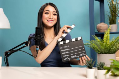 Kadın vlogger kameraya elinde mikrofonla bir masada oturarak bir alkış tahtası sunuyor. Sahne, çevrimiçi incelemeler için video içeriği oluşturma ekipmanı ve atmosferini vurguluyor.