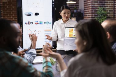 Kadın girişimci, iş analizlerini göstermek için dizüstü bilgisayar kullanarak çalışanlarla mali planlamayı tartışıyor. Akşam toplantısında hazırlık, strateji ve son karar verme süresinin altı çizildi.