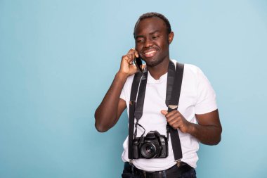 Afro-Amerikalı adam cep telefonuyla konuşurken gülümsüyor, tek eliyle pantolon askısını tutuyor. Boynunda kamera olan neşeli bir fotoğrafçı ayakta duruyor ve telefonda konuşuyor..