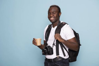 Sırt çantası ve kullan-at bardakla gülümseyen erkek model doğrudan kameraya bakıyor, yeni bir yaratıcı proje için özgüven ve hazırlık yayıyor. Siyah fotoğrafçı elinde teçhizatı ve kahvesiyle.