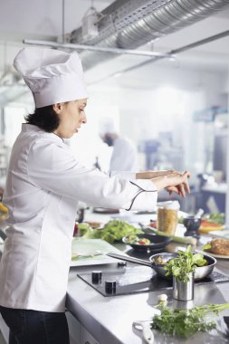 Restoran mutfağındaki genç kadın şef dikkatlice taze otları bir tabağa serpip maydanoz, biberiye ve fesleğenle lezzeti arttırıyor. Taze ürün ve baharatla dolu modern çalışma alanı.