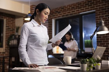 Odaklanmış bir çalışan elinde bir belge okuyor, gece vardiyası sırasında ofis masasında duruyor. Asyalı şirket yöneticisi, geç saatlere kadar çalışıyor, yeni başlayan şirket için bir marka stratejisi planlıyor..