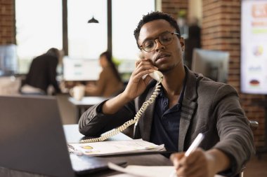 Siyahi erkek yönetici not alırken ev telefonundan iş arkadaşıyla konuşuyor. Elinde kalem olan Afro-Amerikalı bir işadamı, müşterisini dinlerken kağıda yazı yazıyor..