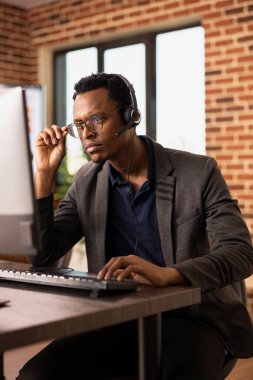 Dalgın siyahi adam gözlüğünü ayarlıyor ve masaüstü bilgisayarında çalışıyor. Müşteri bilgilerine odaklanmış. Kulaklık takan konsantre çağrı merkezi ajanı bilgisayarındaki müşteri destek görevleriyle ilgileniyor..