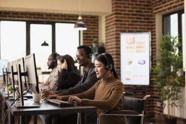 Tekerlekli sandalye kullanan, kablolu kulaklık takan ve ofis masasında özenle çalışan bir kadın. Kulaklıklı Asyalı iş kadını çevrimiçi müşterilerle konuşuyor ve bilgilerini bilgisayara yüklüyor.