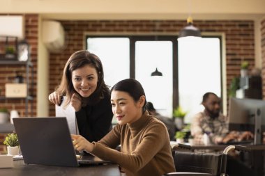 Ofis masasında birlikte çalışan çok ırklı çalışanlar dizüstü bilgisayardaki mali verileri gözden geçiriyorlar. Kafkasyalı yönetici ve Asyalı tekerlekli sandalye kullanıcısının iş büyüme stratejilerini tartıştığı kapsamlı bir çalışma alanı.