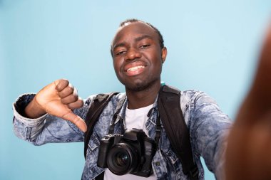 Boynunda profesyonel bir kamera olan ve stüdyoda baş parmağını sallayan mutlu Afro-Amerikan fotoğrafçı. Siyah bir erkek vlogger 'ın POV' u kendisini kaydederken aynı zamanda karşı bir jest yapıyor..