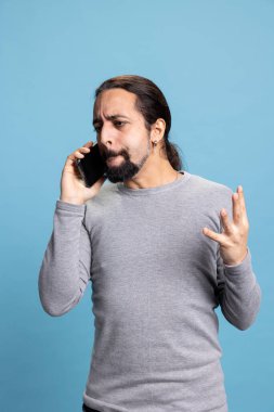 Öfkeli genç bir adam, bir arkadaşıyla yaptığı telefon görüşmesinde sinirlenip sinirleniyor, bağırıyor ve mavi arka plana karşı öfkeli hissediyor. Erkek bir yetişkin sözleriyle sinirli ve agresif..