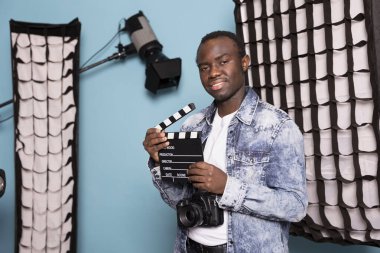 Afrika kökenli Amerikalı film yapımcısı elinde bir klasörle, ışık ekipmanıyla stüdyoda dikiliyor. Boynunda kamera ve film tahtası asılı siyah erkek fotoğrafçının portresi..