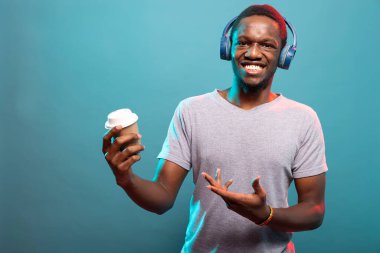 Neşeli genç adam kahve fincanına el sallıyor ve kameraya bakıyor. Siyah erkek portresi sıcak bir içecekle ayakta duruyor ve kablosuz kulaklıklarıyla müziğin keyfini çıkarıyor..