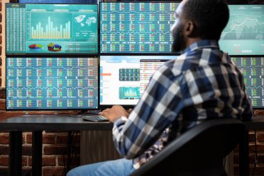 Afrika kökenli Amerikalı Forex yatırımcısı tuğla duvar ofisindeki çok ekran bir kurulumun gerçek zamanlı borsa verilerini takip ediyor. Gece vardiyasında çalışan bir erkek, masa başında oturmuş, ticaret çizelgeleri ve istatistikleri analiz ediyor..