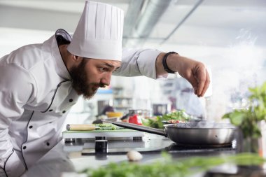Modern bir restoranın mutfağında, beyaz üniformalı şef kaplama bir yemeği baharat ve otlarla süslüyor. Renkli sebzeler, mutfak aletleri ve taze ürünlerle dolu paslanmaz çelik çalışma alanı..