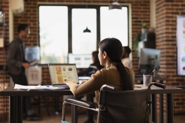 İş eğilimlerini araştırmak için internetli dijital cihaz kullanan pazarlama uzmanı ofis masasında oturuyor. Tekerlekli sandalyedeki kadın yönetici şirket büyüme stratejilerini kişisel bilgisayarında analiz ediyor..