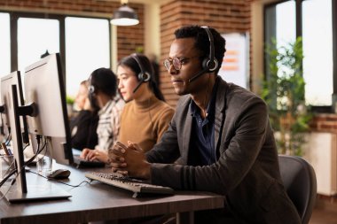 Tuğla duvar ofisindeki bilgisayarında iş analizlerine konsantre olan ciddi bir erkek ofis çalışanı. Odaklanmış, elleri bağlı, kulaklık takan ve masaüstü monitörüne bakan siyah bir işadamı.
