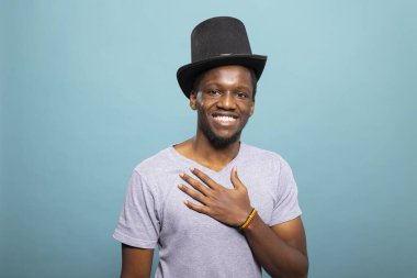 Silindir şapka takan ve eli göğsüne yerleştirilmiş genç bir beyefendi izole bir arka plana karşı duruyor. Siyah başlıklı, kameraya gülümseyen Afrikalı Amerikalı adamın portresi..