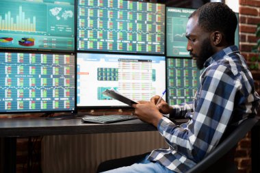 Afro-Amerikan yönetici, şirket koruma fonu için güncellemeleri kaydediyor. Erkek profesyonel not alıyor, ofisteki hisse senedi ticaretinin risk analizi üzerinde çalışıyor..