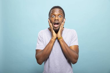 Şok olmuş bakışlı, iki eliyle yüzünü tutan genç bir Afro-Amerikalı. Siyah erkek, geniş gözlü ve ürkmüş bir ifadeyle, izole bir arkaplana kurulmuş..