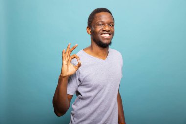 Siyah erkek, parmaklarıyla onay jesti yaparken kameraya gülümsüyor. Gülümseyen Afro-Amerikan adam el işareti yapıyor, anlaşma ve memnuniyeti ifade ediyor..