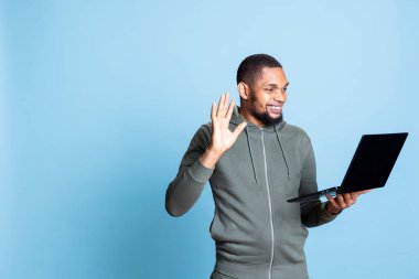 Video konferans sırasında arkadaşlarıyla sohbet eden Afrikalı Amerikalı bir adam mavi arka planda duruyor. Video kameraya el sallayan genç bir yetişkin arkadaşlarıyla uzaktan tanışıyor..