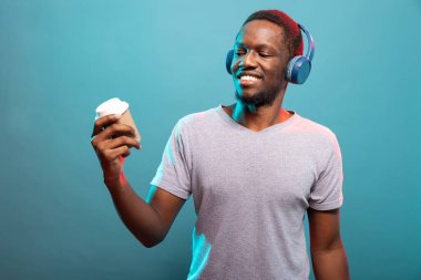 Günlük giysiler içinde gülümseyen bir adam, tek kullanımlık bardağına bakıyor ve izole bir arka planda müzik dinliyor. Kulaklık takan ve elinde kahveyle dikilen Afrikalı bir Amerikalı..
