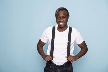 Afro-Amerikalı bir adam, sıradan giyinmiş, askılarıyla stüdyoda poz veriyor ve klasik bir cazibe katıyor. Siyah erkek model izole edilmiş, kendinden emin bir şekilde duruyor..