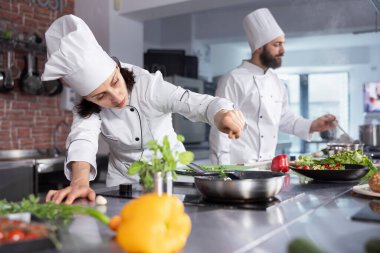 Mutfakta, kadın aşçı taze otlar ekleyerek, fesleğen ve diğer baharatlarla tatları dengeleyerek gurme yemeğini mükemmelleştiriyor. Mutfak tezgahında sebze ve yemek hazırlamak için gerekli malzemeler var..
