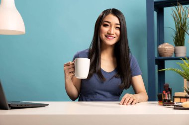 Mavi arka plandaki canlı yayın sırasında çevrimiçi izleyicileriyle konuşurken elinde bir fincan kahve tutan güzel bir kadın. Gülümseyen genç bir vlogger hayranları için bir video kaydediyor.