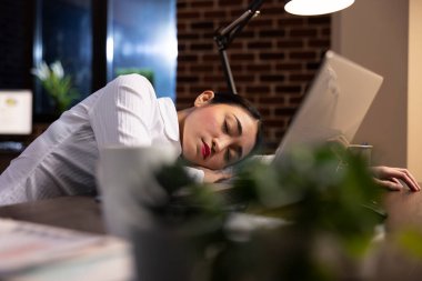 Gece vardiyasından yorgun düşen genç iş kadını, sessiz bir ofiste dizüstü bilgisayarının yanında uyuyor. Asyalı kadın yöneticinin başı masanın üzerinde, etrafı evrak işleri ve dijital cihazlarla çevrili..
