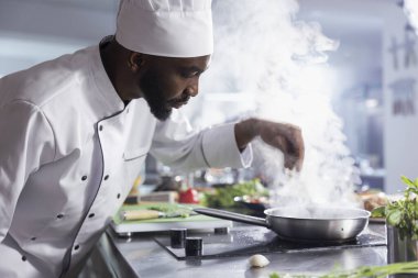 Bir mutfakta, Afro-Amerikan şef tabağa taze ot yaprakları ekliyor. Erkek uzman lezzetli bir yemek yapmak için çiğ sebze, baharat ve gerekli mutfak malzemelerini kullanır..