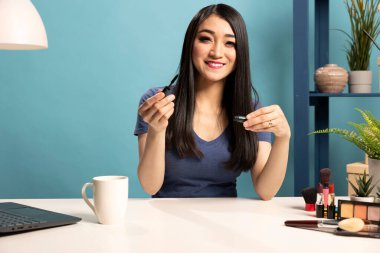Elinde rimel olan Asyalı bir makyöz internetteki seyircileri için özel ders videosu çekiyor. Kadın vlogger masada oturuyor ve hayranlarına güzellik rutinini gösteriyor..