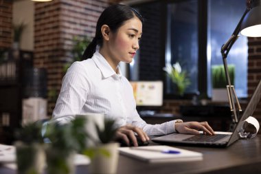 Meslek ortamında genç bir iş kadını, masasında oturuyor, proje sunumu için dizüstü bilgisayar kullanıyor. Modern tuğla duvar tasarımı ofise şık ve üretken bir atmosfer katıyor..