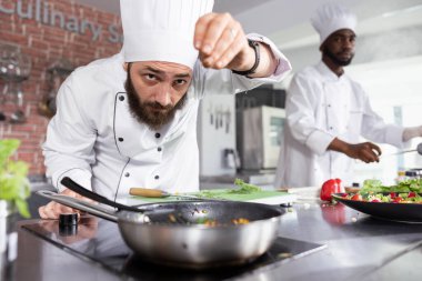 Ticari mutfakta aşçılık uzmanı taze fesleğen ve maydanozlu bir yemek hazırlıyor, iyi yemek ve yemek hazırlama konusunda gastronomi uzmanlığı gösteriyor. Genç adam şef biberiye yaprakları serpiyor..