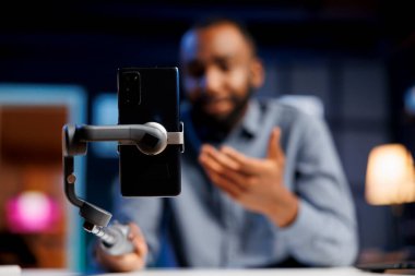 İçerik yaratıcısı tarafından canlı yayın yapmak için kullanılan selfie çubuğuna bağlı cep telefonuna odaklan. Canlı yayın yapmak için cep telefonu kullanan coşkulu internet flaması, bulanık arkaplan