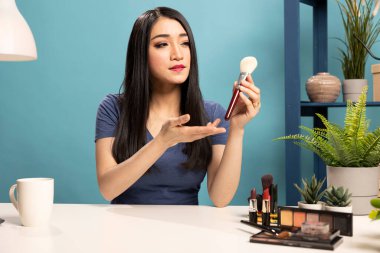 Güzellik vlogcusu elinde fırça tutarken makyaj ipuçlarını açıklıyor. Asyalı kadın masasında oturmuş, kozmetik aletine el kol hareketi yapıyor, özel bir video hazırlıyor ve online takipçileriyle bağlantı kuruyor..