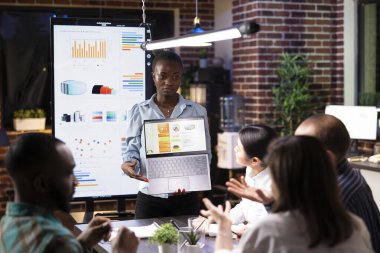 Odaklanmış kadın yönetici, gece vardiyası toplantısında iş analizlerini laptopta sunar. Modern çalışma alanındaki dijital cihazlar üzerindeki finansal tabloları analiz ederek, çok ırklı iş arkadaşları tartışmalara giriyorlar..