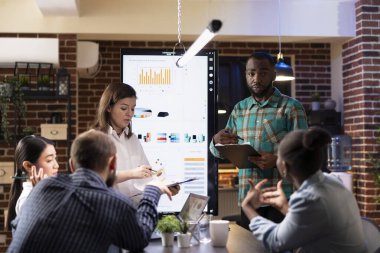 Tuğla duvar ofisinde çalışan çok ırklı çalışanlar, yaklaşan iş sunumuna hazırlanmak için pazarlama eğilimlerini ve dijital ekrandaki finansal grafikleri gözden geçiriyorlar. Gece yarısı toplantısında takım çalışması.