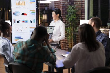 Kadın girişimci finansal tabloları analiz eder, iş arkadaşlarıyla pazarlama stratejisini neşeyle tartışır. Profesyonel Asyalı kadın dizüstü bilgisayar tutuyor, grup tartışması sırasında gülüyor..