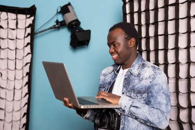 Elinde dizüstü bilgisayarıyla gülümseyen siyah fotoğrafçı resimlerini kontrol ediyor. Mutlu Afrikalı Amerikalı adam kişisel bilgisayarını tutuyor ve fotoshoot seansından sonra resimlerini inceliyor..