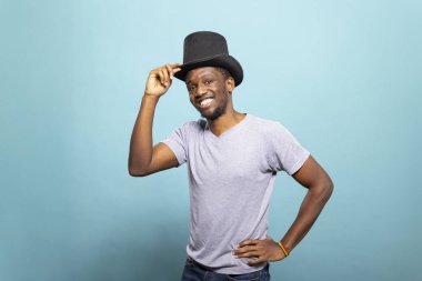 Genç siyah adam silindir şapkasını düzeltiyor ve mavi arka planda kameraya poz veriyor. Mutlu erkek illüzyonist klasik bir sihirbaz elbisesi giyiyor ve eli belinde duruyor..
