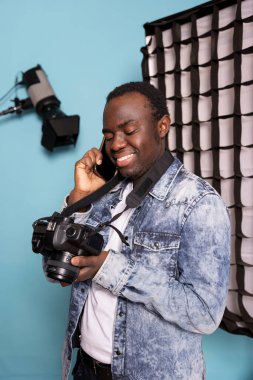 Afrikalı Amerikalı bir adam kamera ekranına bakıyor, resimlere bakıyor ve stüdyodaki bir görüşmede müşteriyle konuşuyor. Siyahi erkek fotoğrafçı cep telefonunu kulağına dayamış, mutlu bir şekilde fotoğraf galerisine bakıyor..