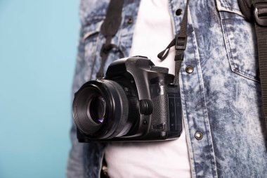 İzole edilmiş arka planda kot ceket ve beyaz gömlek giyen bir kişinin üzerinden sarkan DSLR 'nin yakın çekimi. Bir insanın vücuduna yerleştirilmiş profesyonel bir kameraya seçici odaklanma.