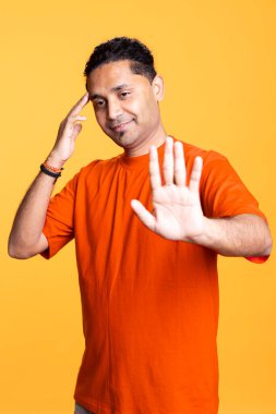 Hintli arkadaş canlısı, erkek topuzlu, elini kaldıran ve el sallayan turuncu arka planda arkadaşlarını selamlayan bir adam. Günlük giysiler içinde genç bir yetişkin, keyifli bir adam..