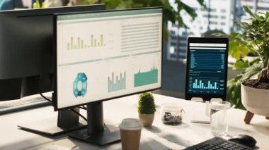 Parlak ofis ortamındaki cihazlarda analitik veri göstergelerine sahip modern çalışma alanı. Finansal istatistik grafikleri saksı bitkilerinin yanında olan masadaki bilgisayar ekranı, dümen aşağı