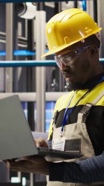 Akıllı fabrikada çalışan dikey video uzmanı güvenilirliğini sağlamak için otomatik sistemleri test ediyor. Endüstri 4.0 'daki Afrikalı Amerikalı adam not defterini kullanarak darboğazları tespit ediyor ve optimize ediyor.