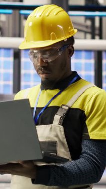 Güneş panelleri fabrikasındaki gülümseyen teknisyenin dikey video portresi robot montaj hatlarını denetliyor ve kalite kontrolü sağlıyor. Endüstriyel fabrikadaki Afrikalı Amerikalı adam otomatik taşıyıcıyı izliyor