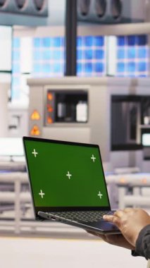 Endüstriyel fabrikadaki dikey video teknisyeni ekipman hatalarını teşhis etmek için yeşil ekran laptop kullanıyor. Montaj hatlarındaki tamir görevlerini koordine etmek için krom anahtar model dizüstü bilgisayar kullanan bir fabrika uzmanı