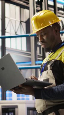 Dikey video Akıllı güneş panelleri fabrika teknisyeni dizüstü bilgisayarını taşıyıcı bantları denetlemek için kullanıyor. Endüstrideki Afrikalı Amerikalı adam 4.0 fabrika makinelerinin en uygun verimlilikte çalıştığından emin oluyor.