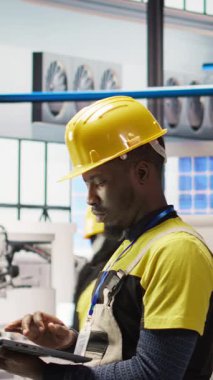 Akıllı fabrikadaki dikey video teknisyeni sistem performans ölçümlerine bakmak için defter kullanıyor. IoT sensörleri aracılığıyla enerji üretimini takip etmek için dizüstü bilgisayar kullanan 4.0 sanayiindeki Afrikalı Amerikalı adam.